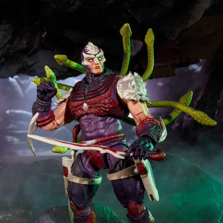 Hasbro product photograph of the Nemesis Immortal figure posed in a dark cave diorama wearing the alternate unmasked head with pale skin and purple markings, with the green segmented tentacle attachment fanned out behind his shoulders and a curved sword held across his body