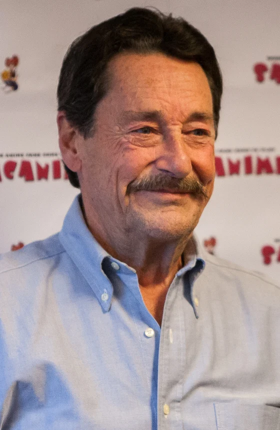 Peter Cullen in 2015, smiling in a light blue shirt in front of a convention backdrop.